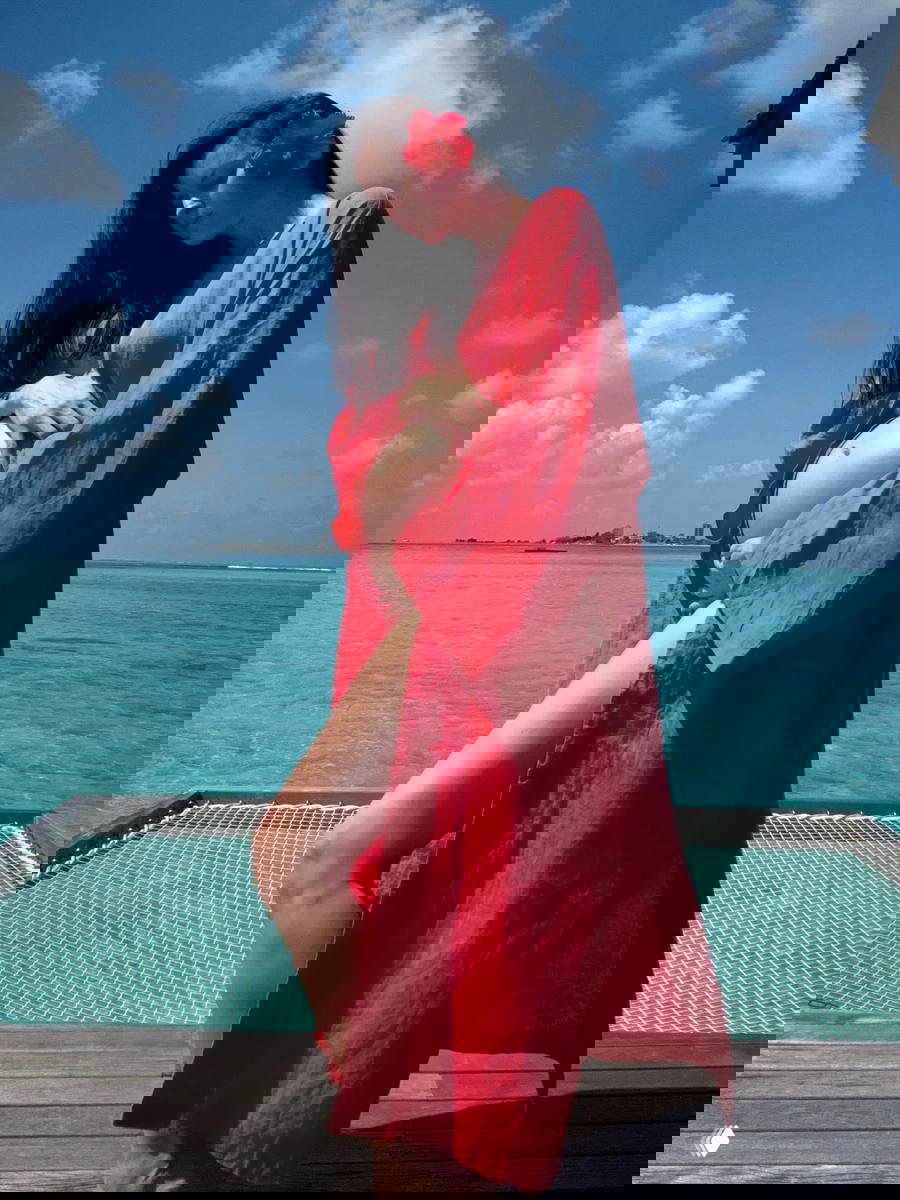 Марина Ворожищева на отдыхе у моря в красном лёгком халате, держит беременный живот, цветок в волосах на фоне бирюзовой воды