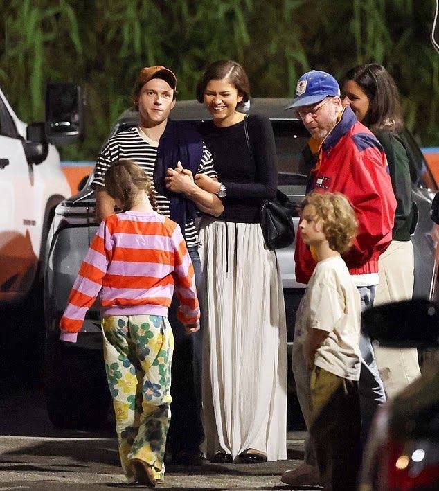 Широкий папарацци‑кадр: пара у машины, несколько детей и прохожих вокруг, ночная уличная сцена с мягким освещением.