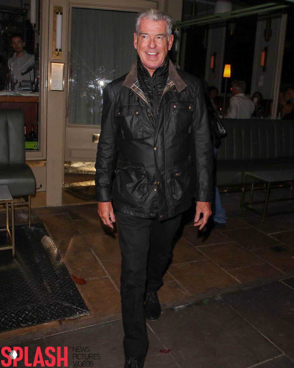 Пирс Броснан улыбается фотокамерам у входа в ресторан в Лондоне: кадр показывает актёра в чёрной куртке и тёплой атмосфере вечера.