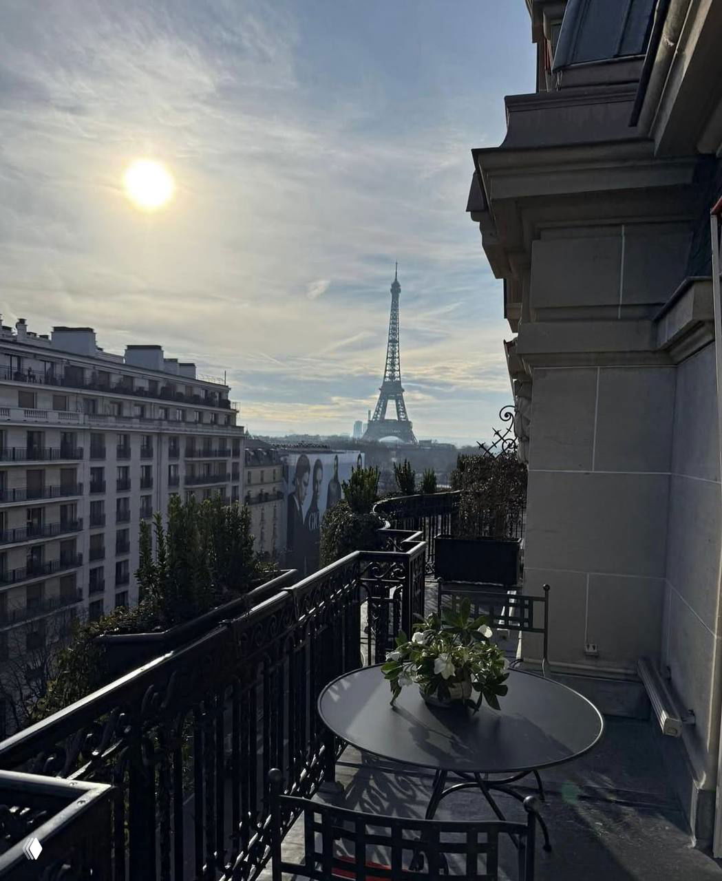 Вид с парижского балкона на Эйфелеву башню: столик на террасе, утреннее солнце и ряд городских зданий вокруг.