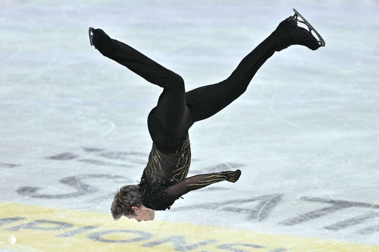Илья Малинин в прыжке, тело в воздухе над льдом в чёрном костюме — динамичный кадр произвольной программы на чемпионате мира