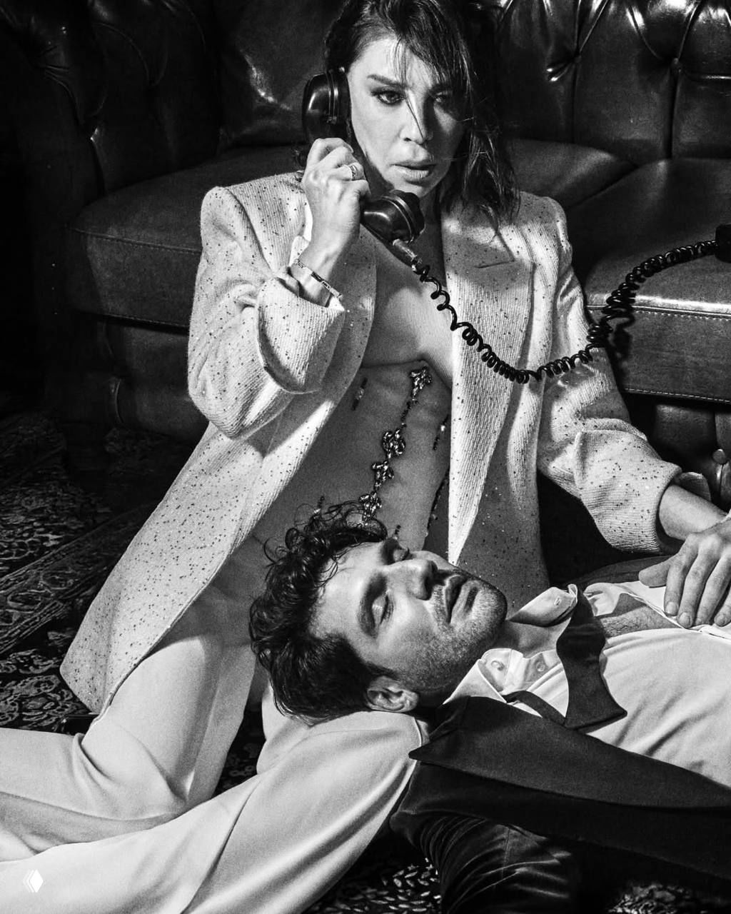 Драматичный кадр: Каан лежит на коленях Небахат, актриса держит телефон — кинематографичный чёрно‑белый образ с театральной интонацией.