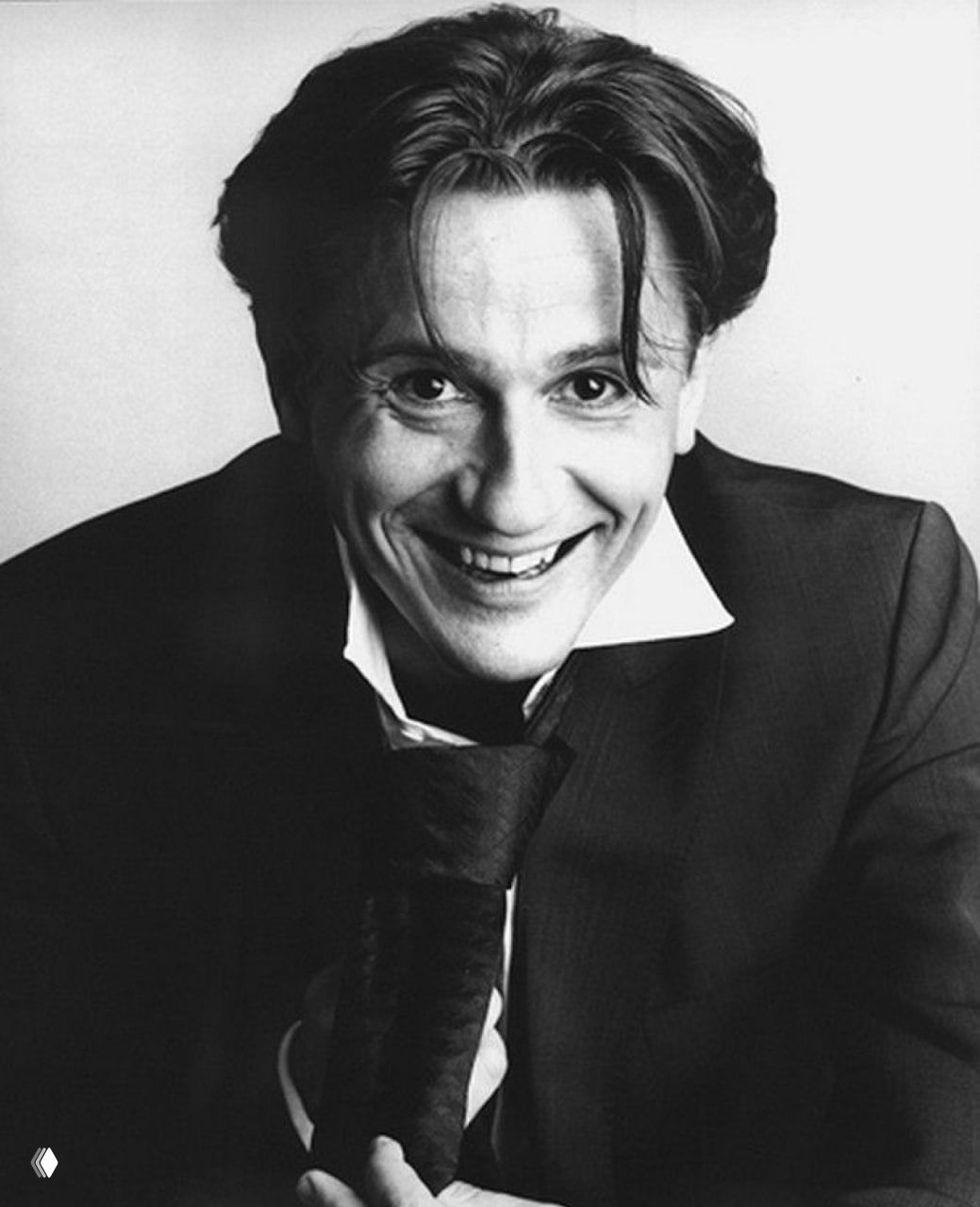 Чёрно‑белая архивная фотография молодого мужчины в костюме, крупная улыбка, артистичное выражение лица и лёгкая небрежность прически.