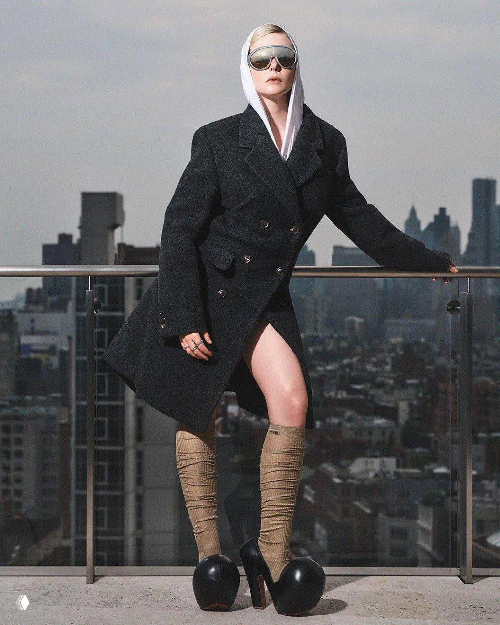 Фотомодель в пальто и капюшоне с оттенком спорт-шика стоит на балконе с неброским городским фоном — сочетание уличной моды и высокой редакционной эстетики.