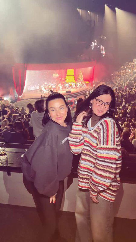 Две женщины в зрительном зале на фоне сцены с подсветкой; снимок сделан из ложи во время концерта, видна сцена и публика в глубине кадра.