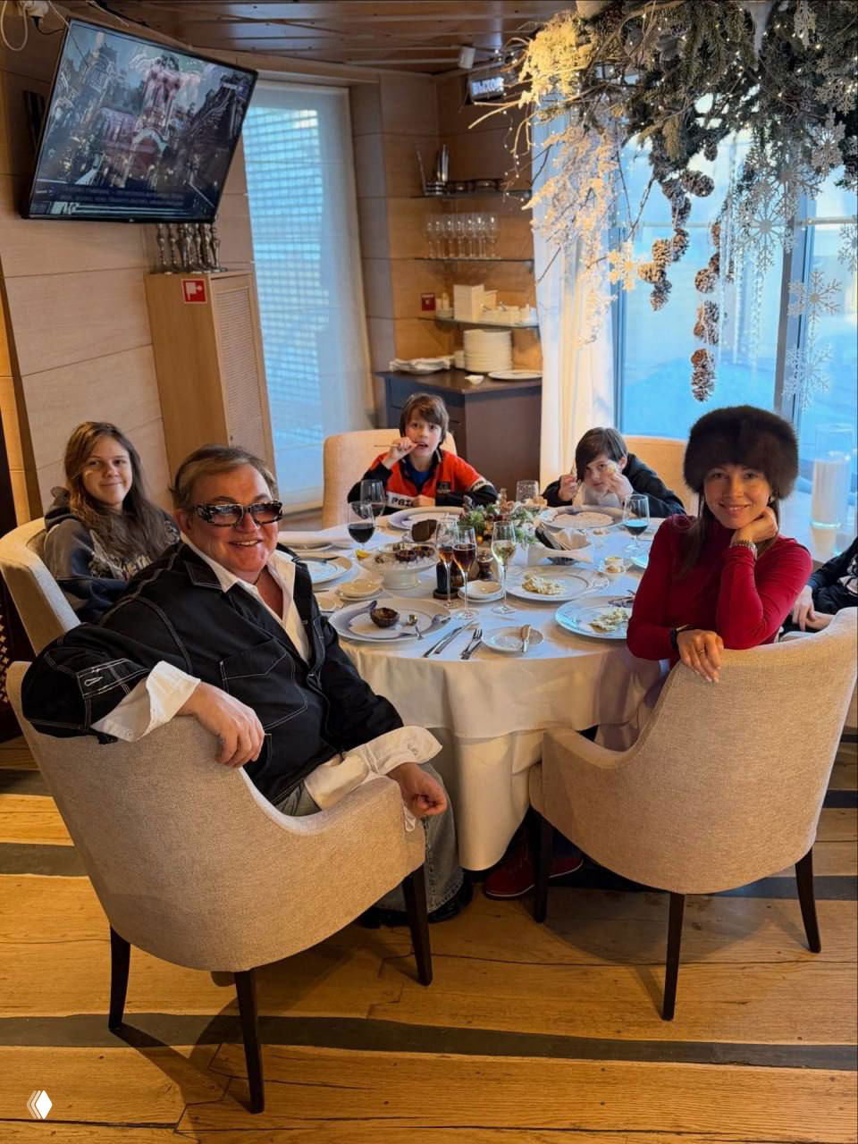 Семья и дети сидят за богато накрытым праздничным столом в ресторане; видны блюда и зимние украшения интерьера.