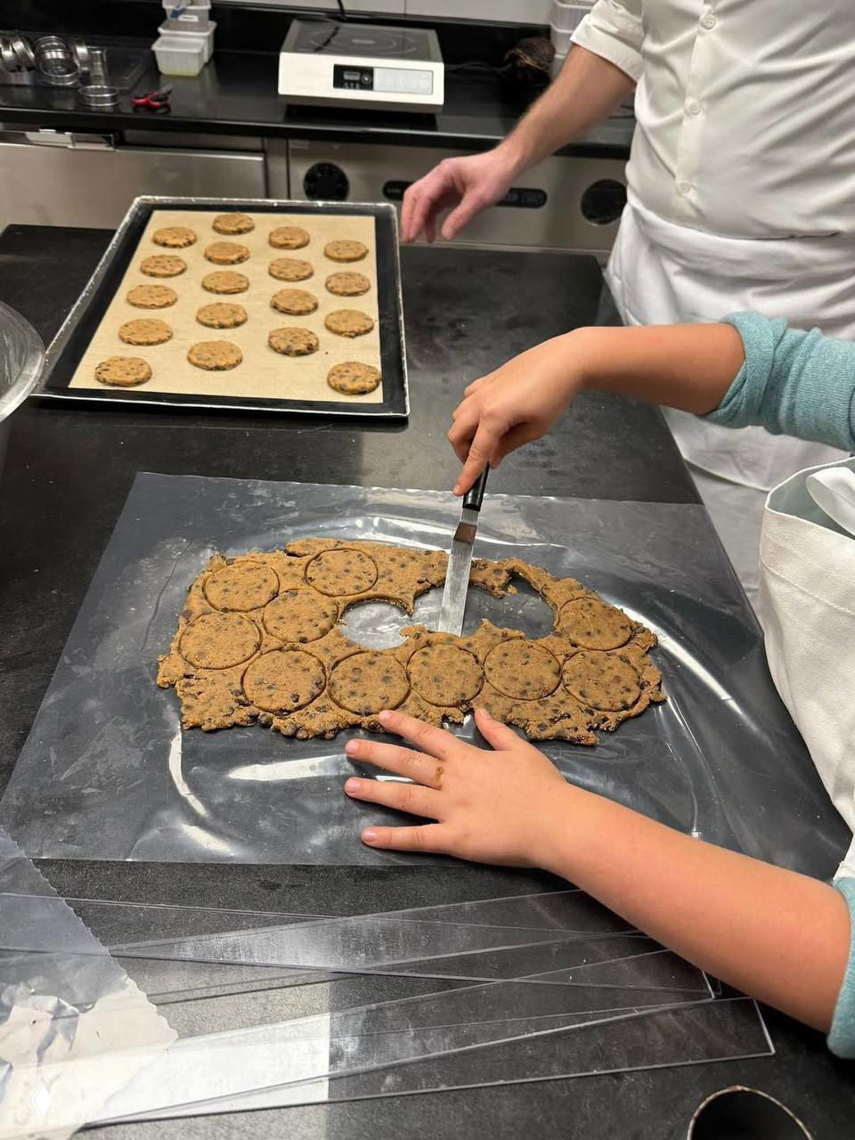 Кулинарный кадр в пекарне: раскатанное тесто, вырезанные кружки печенья на столе и руки ребенка, помогающего на кухне.