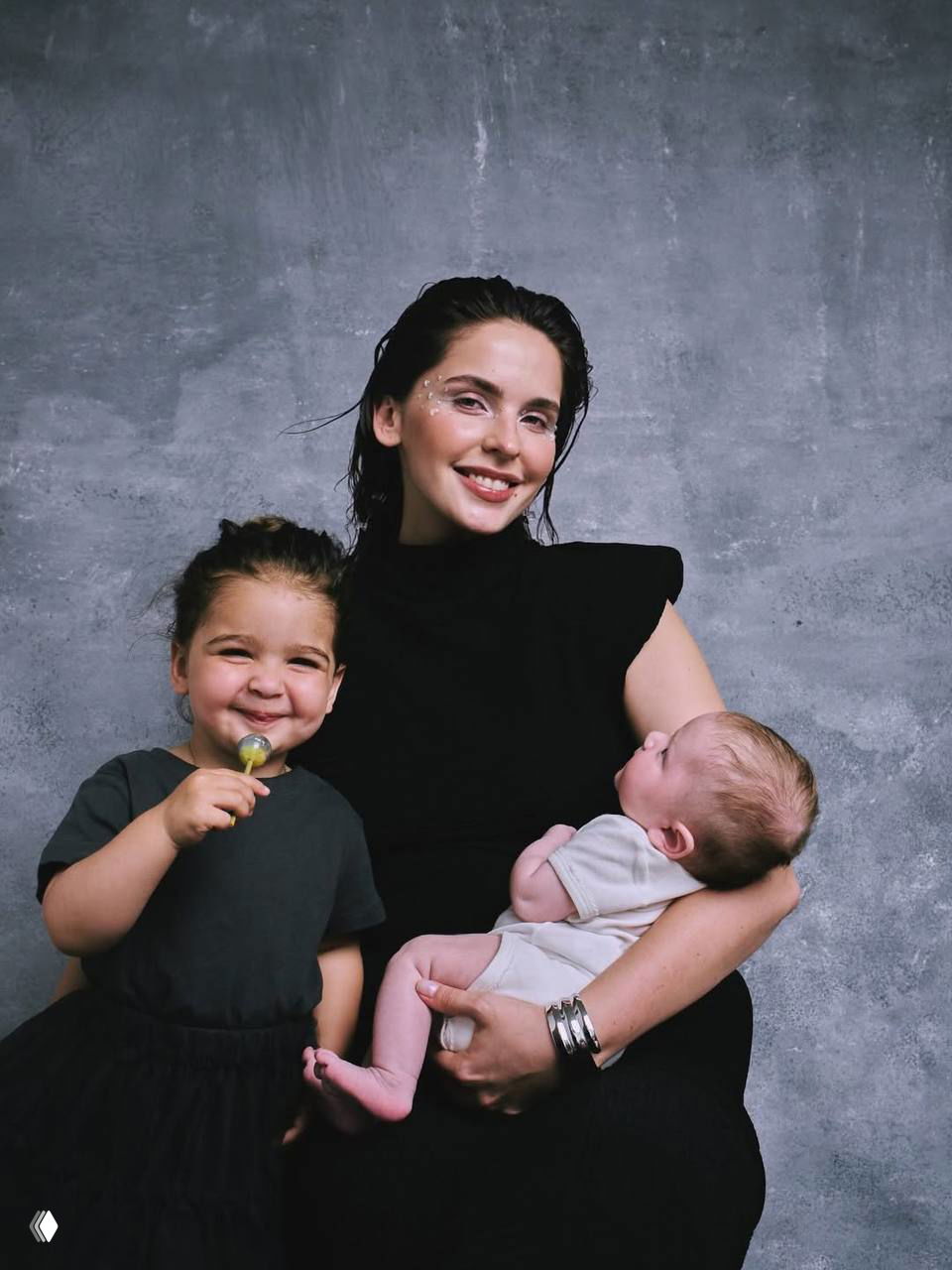 Портрет женщины с тёплой улыбкой, она держит на руках младенца, рядом стоит маленький ребёнок — студийный снимок с нейтральным фоном и нежной атмосферой