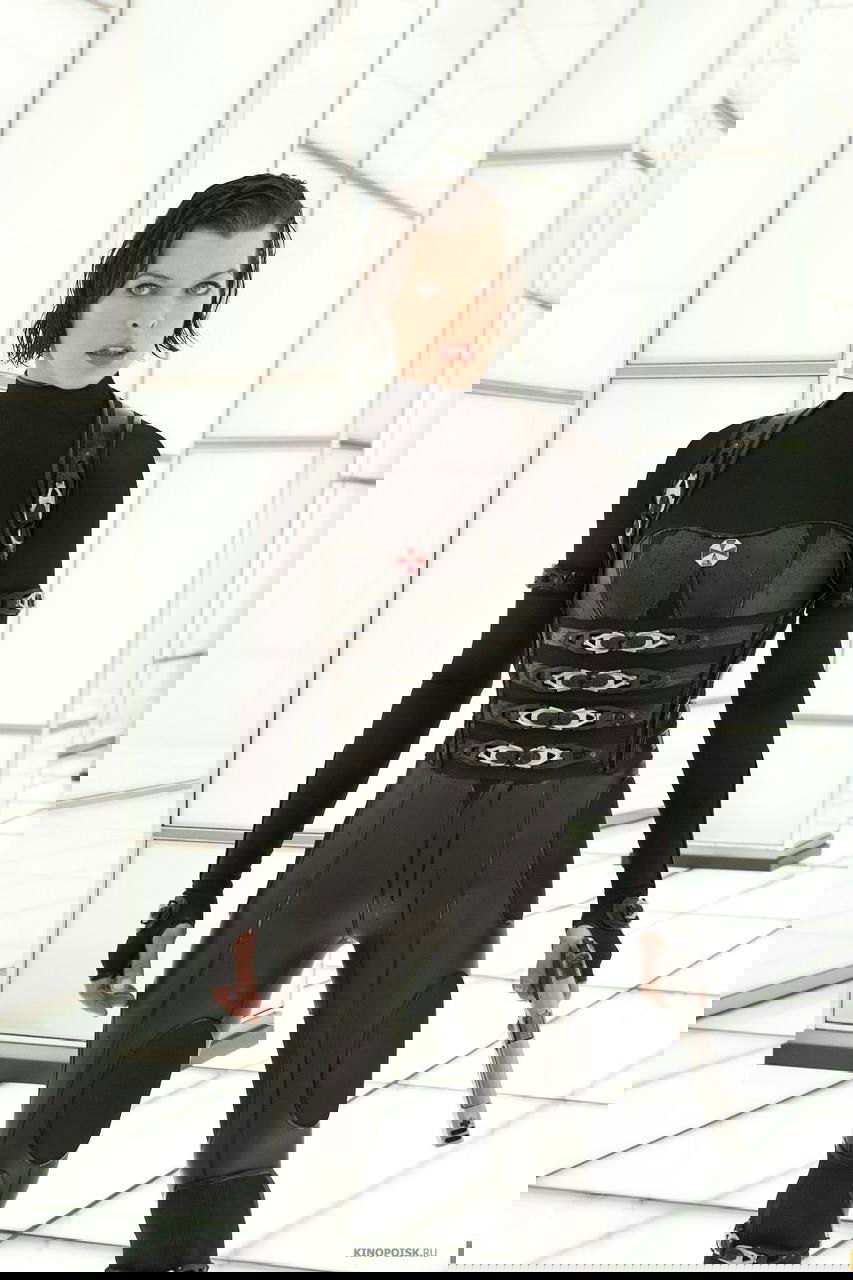 Полнофигурный кадр актрисы в чёрном облегающем костюме: сцена с минималистичным белым фоном, атрибутика боевого образа.