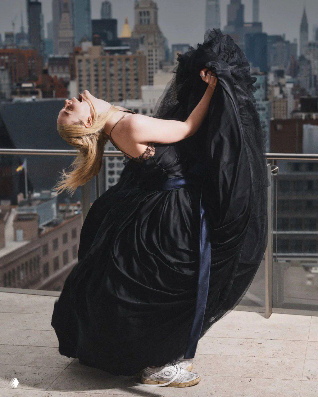 Динамичный кадр женщины-модели в развевающемся черном платье на балконе, город за спиной — сочетание драмы и уличной эстетики в редакционной съемке.