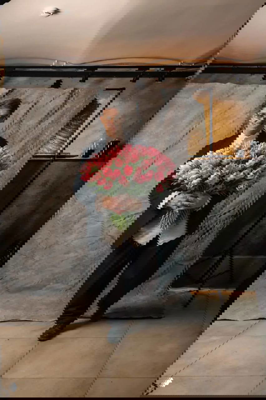 Мужчина в строгом костюме позирует в фотостудии, держит пышный букет розовых тюльпанов на фоне серой текстурной простыни и со светотеневым оформлением