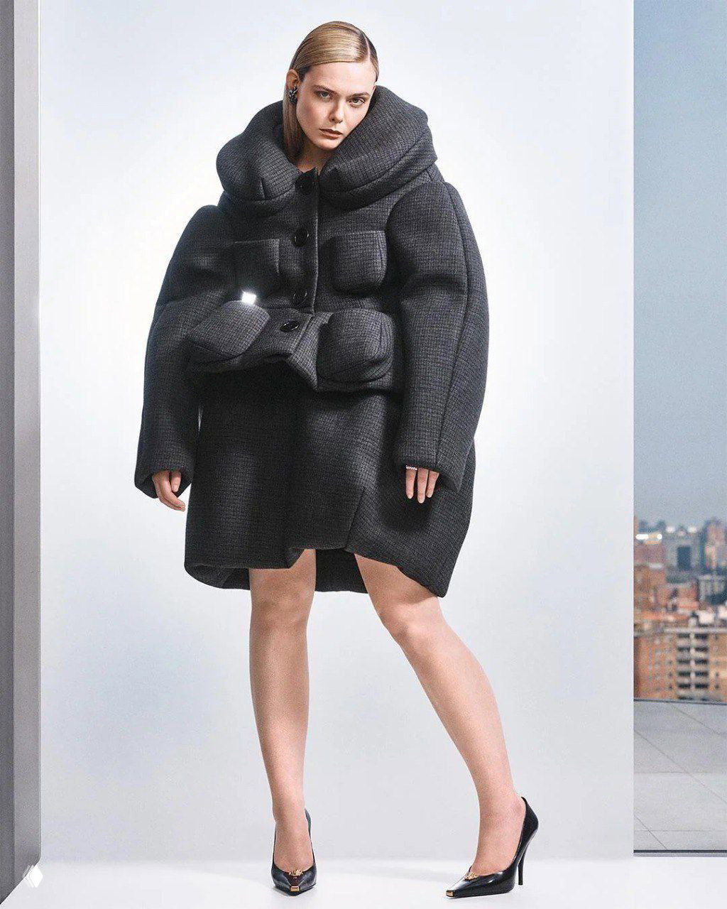 Полноформатный портрет в объемном стеганом пальто на нейтральном фоне с намеком на городской пейзаж — стилизованный образ из фотосета для журнала.