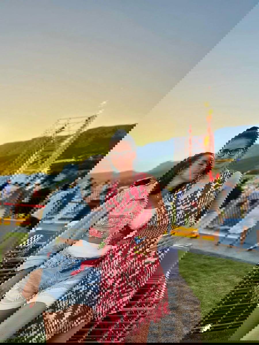 Две молодые женщины на открытом летнем мероприятии у сцены на закате; на заднем плане сцена, публика и тёплая атмосфера уличного фестиваля.