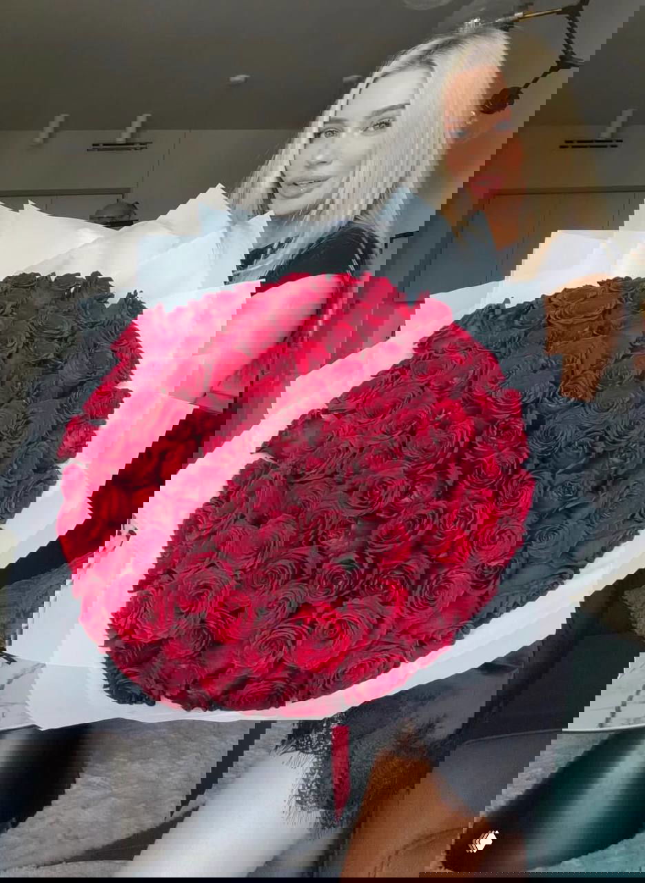 Женщина держит огромный букет насыщенно‑красных роз в белой бумажной упаковке, интерьер квартиры виден на заднем плане, акцент на размере букета