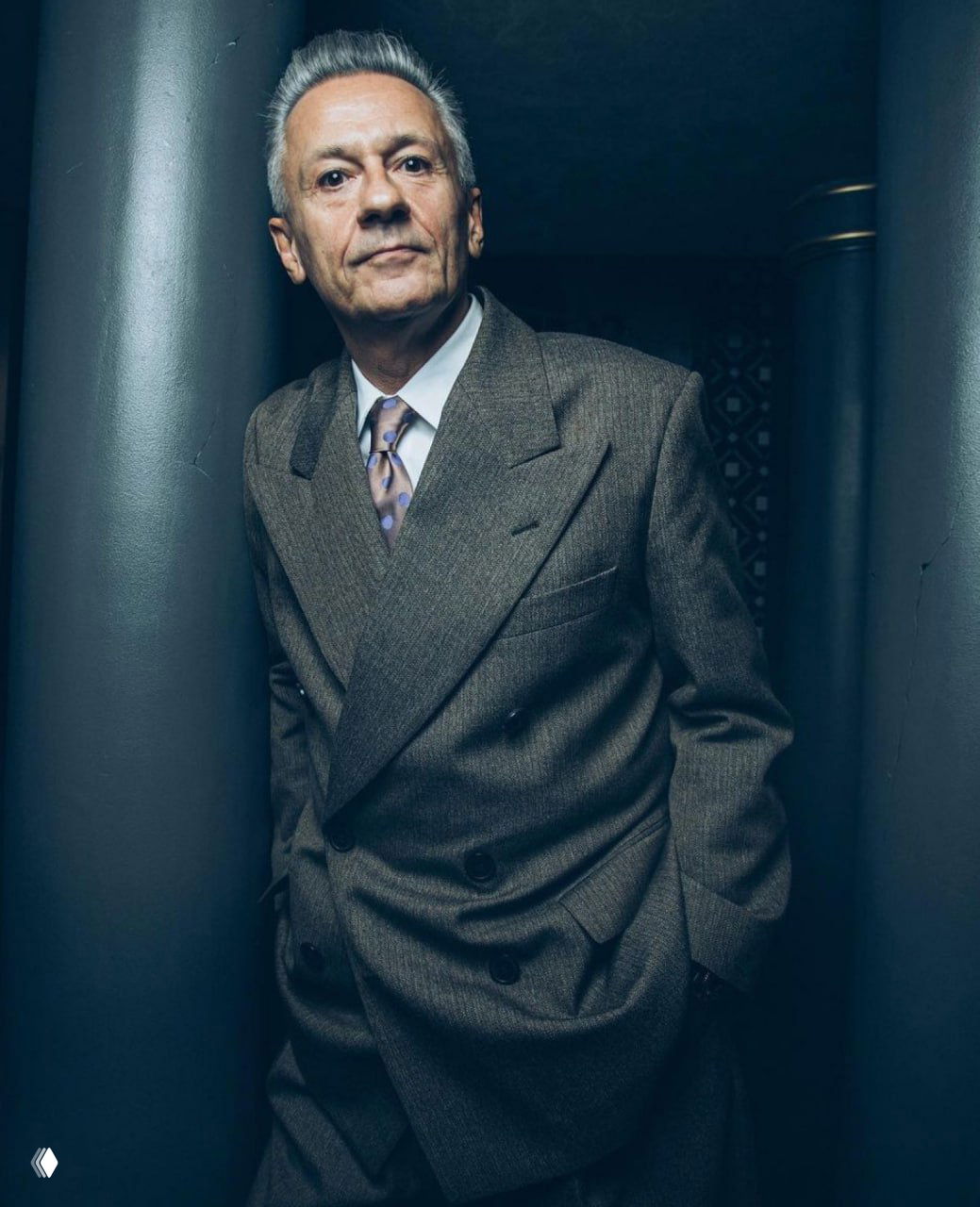 Фотография зрелого мужчины в классическом двубортном костюме, строгая поза в узком коридоре, холодная тональность и подчёркнутая фактура ткани.