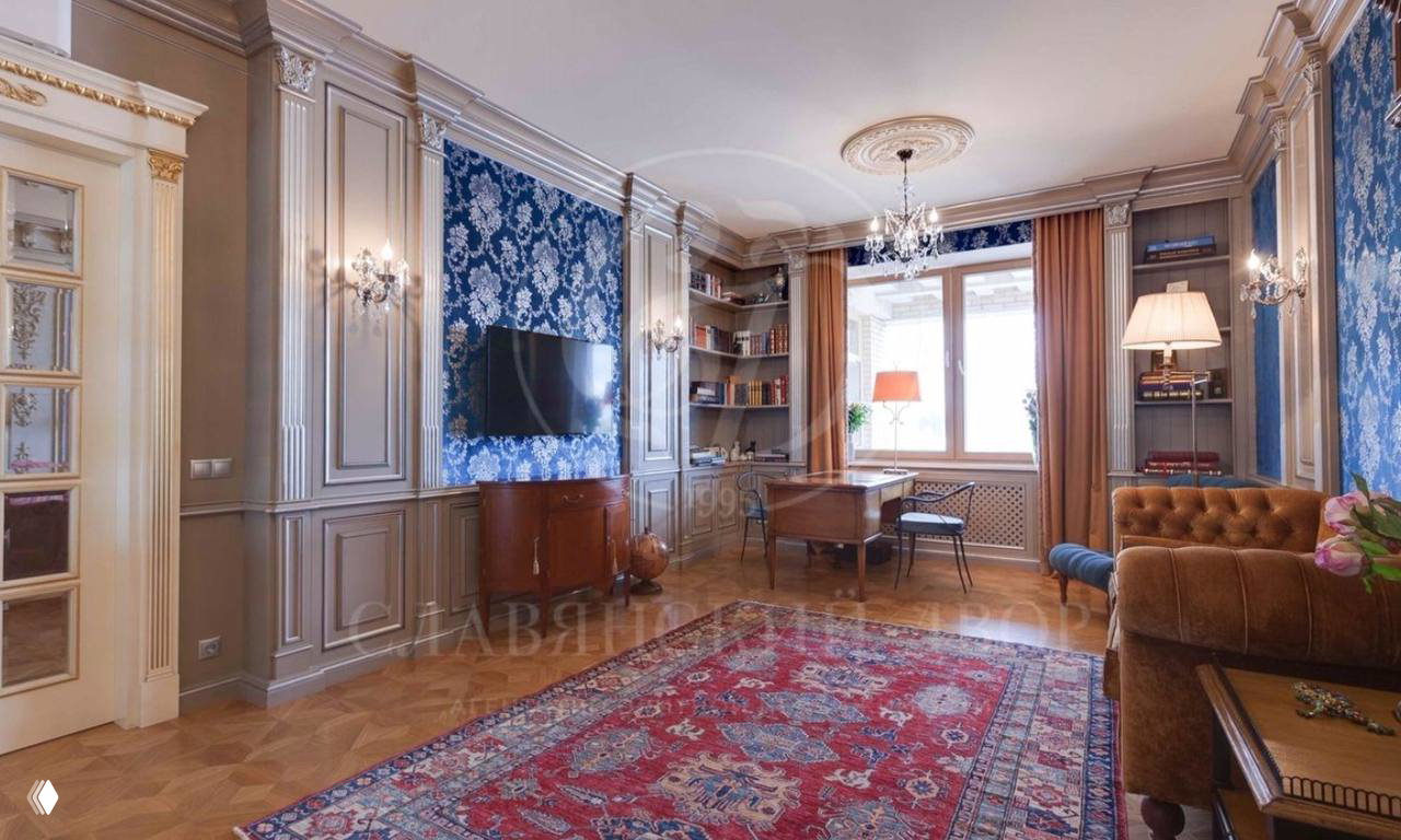 Элегантная гостиная с резными стеновыми панелями, синими обоями, книжными полками, столом у окна и декоративным ковром на паркете.