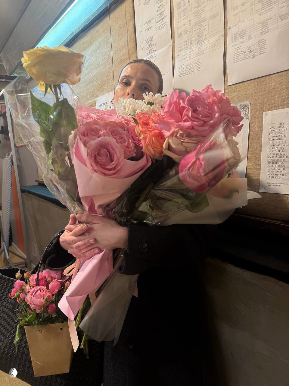 Актриса за кулисами театра держит большой букет розовых и жёлтых роз, вокруг видна гримёрная комната, афиши и реквизит.