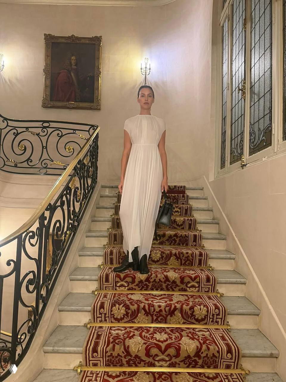 Элегантный кадр на лестнице дворца: Ирина Шейк в длинном белом платье стоит на ковровой дорожке среди классического интерьера и витражных окон.