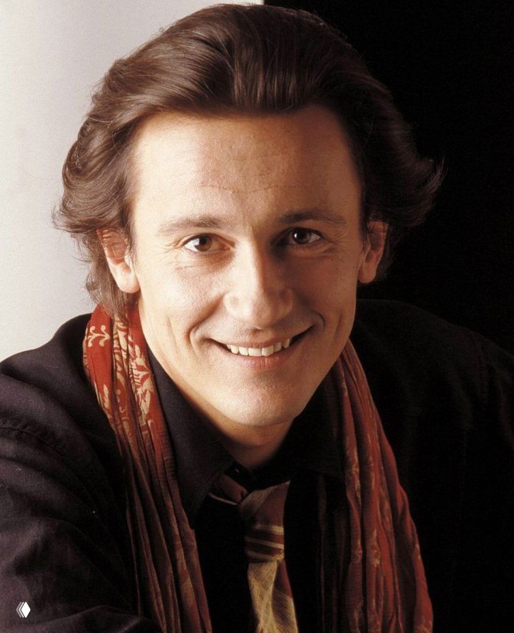 Крупный план улыбающегося молодого актёра с шарфом, тёплые тона, естественное выражение лица и мягкое освещение студии.