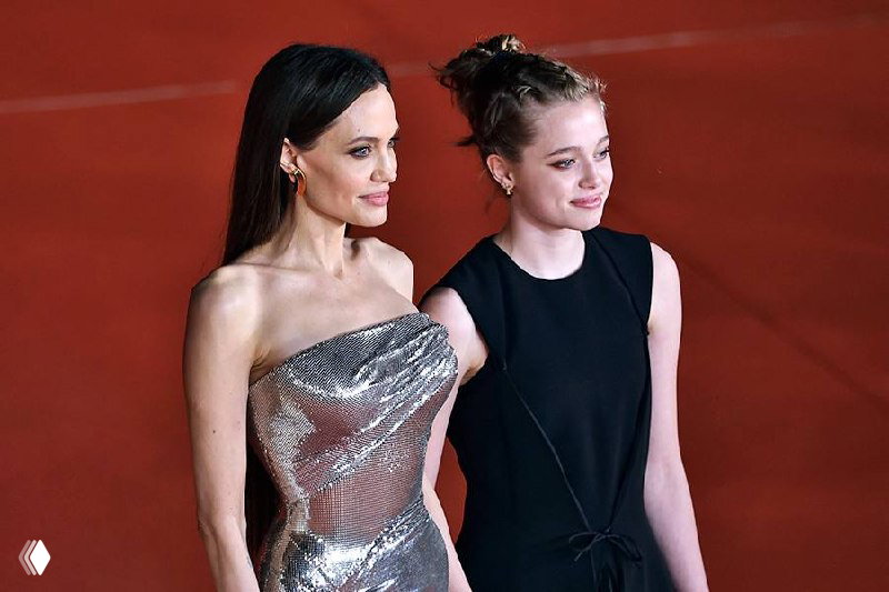 Та же пара в вечерних нарядах проходит по красной дорожке в общем плане; камера снимает сбоку, на заднем плане зрители.