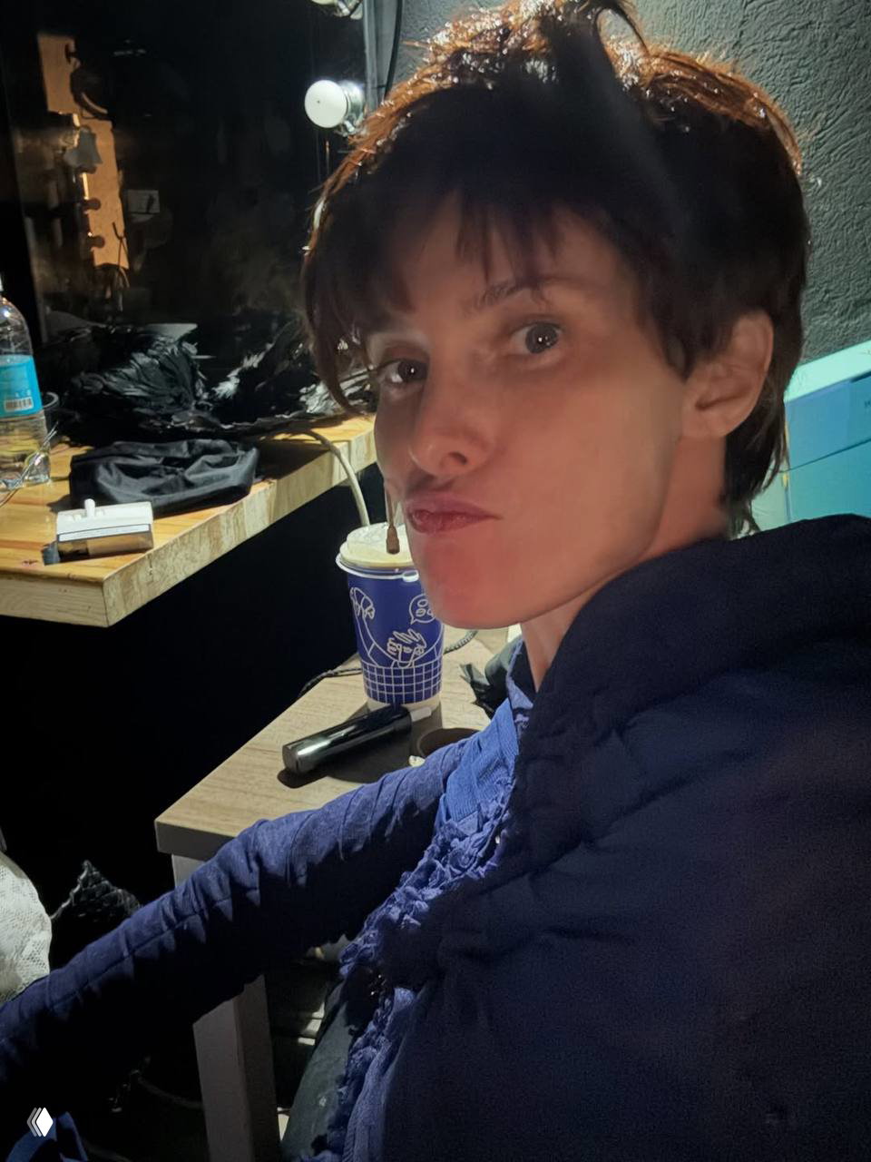 Крупный портрет актрисы в гримёрной зоне: селфи у рабочего стола с чашкой кофе, на заднем плане реквизит и осветительные приборы.