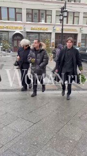 Группа людей идёт по тротуару с цветами в руках возле театра; прохожие и фасады зданий на заднем плане, на кадре водяной знак WOMAN.ru