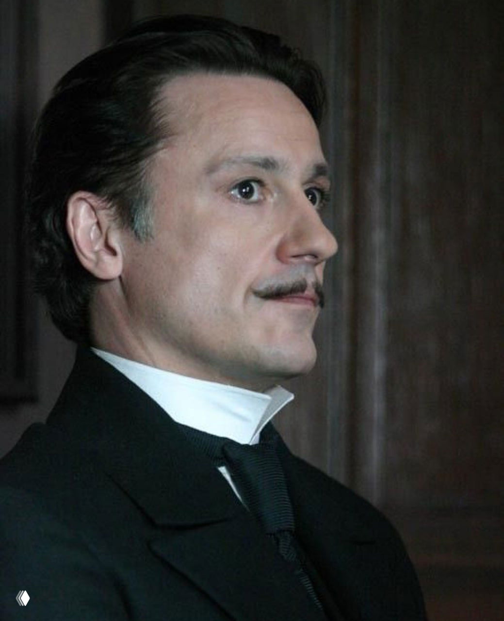 Профильный кадр в театральной сцене: мужчина в строгом костюме, подчёркнутый профиль и фокус на взгляде, драматическая постановка света.