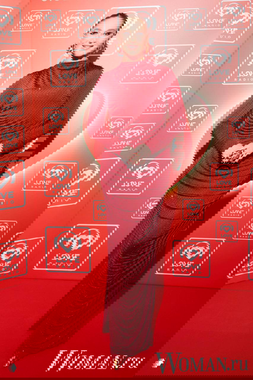 Женщина в бордовом длинном платье с высокой горловиной и блеском стоит на красной дорожке у декорации Big Love Show.