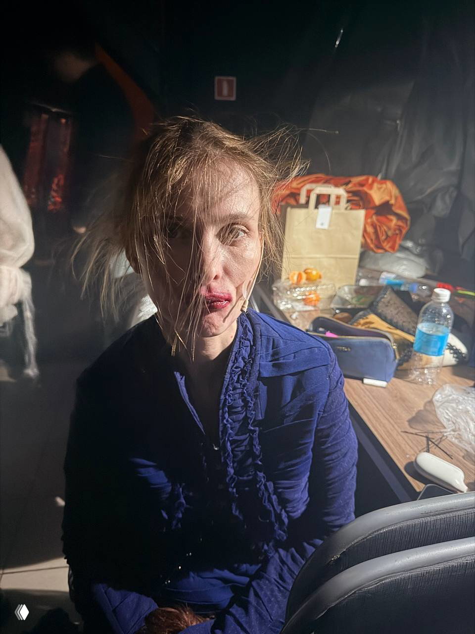 Актриса с небрежной причёской и гримом сидит в гримёрной комнате среди реквизита, сумок и бутылок воды на столе за ней.