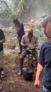 Дуэйн Джонсон на съёмочной площадке в лесу: он сидит с тарелкой и большой ложкой, рядом находятся члены съёмочной группы и реквизит.