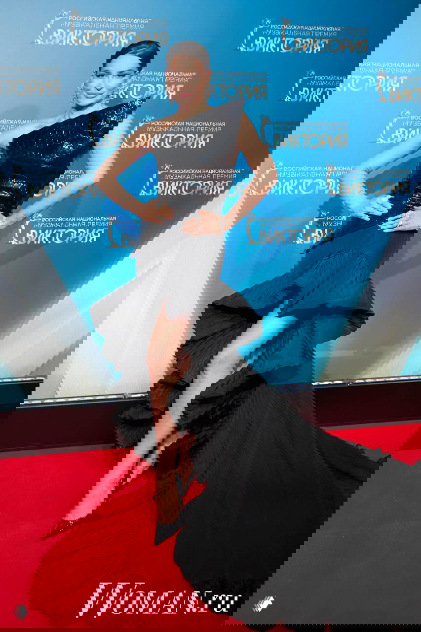 Знаменитость в асимметричном чёрном платье с высокой волнистой юбкой и глубоким разрезом позирует на красной дорожке «Виктория-2026».