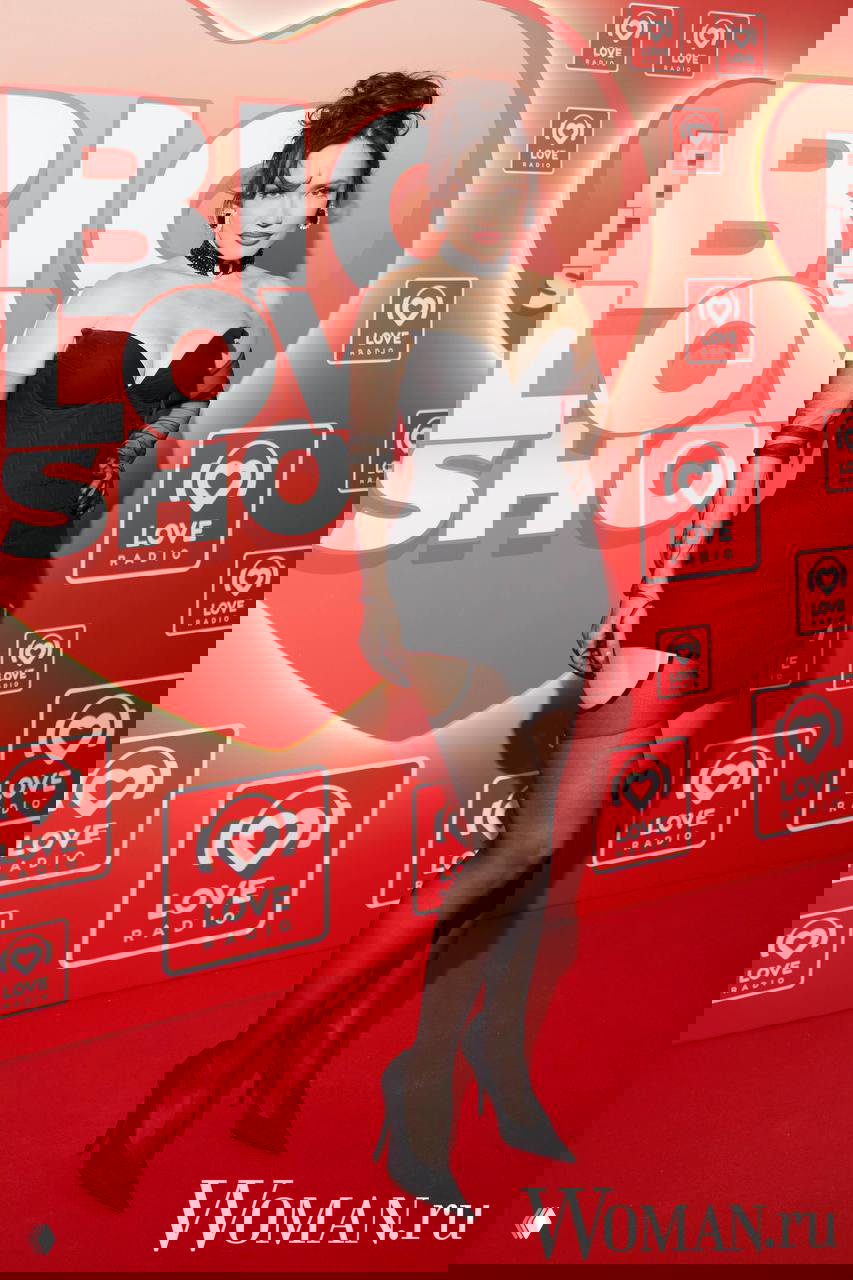 Женщина в чёрном платье-корсете с чулками и чокером позирует на красной ковровой дорожке у стенда Love Radio.