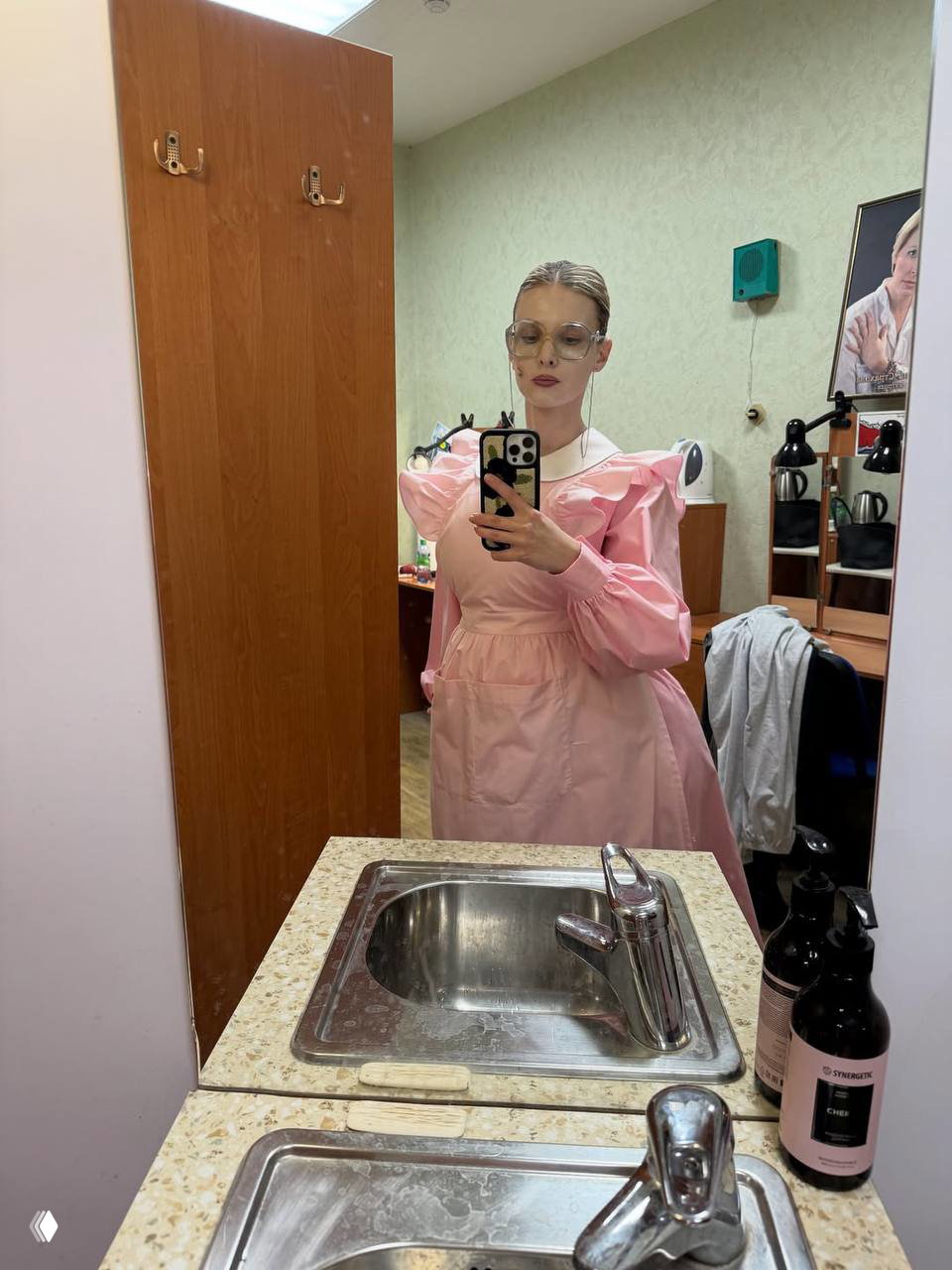 Зеркальное селфи актрисы в розовом платье в гримёрной комнате; на переднем плане видна раковина и флаконы с косметикой.