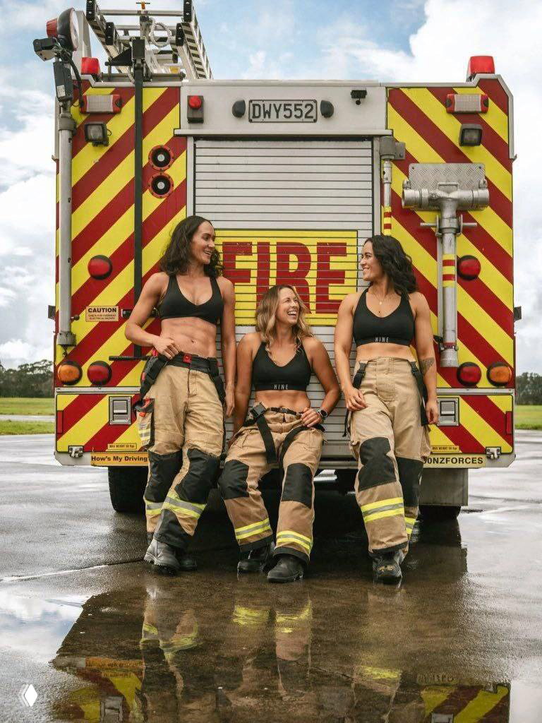 Три женщины‑пожарные в рабочей форме стоят у задней части пожарной машины с яркими диагональными полосами; видна влажная отражённая поверхность.