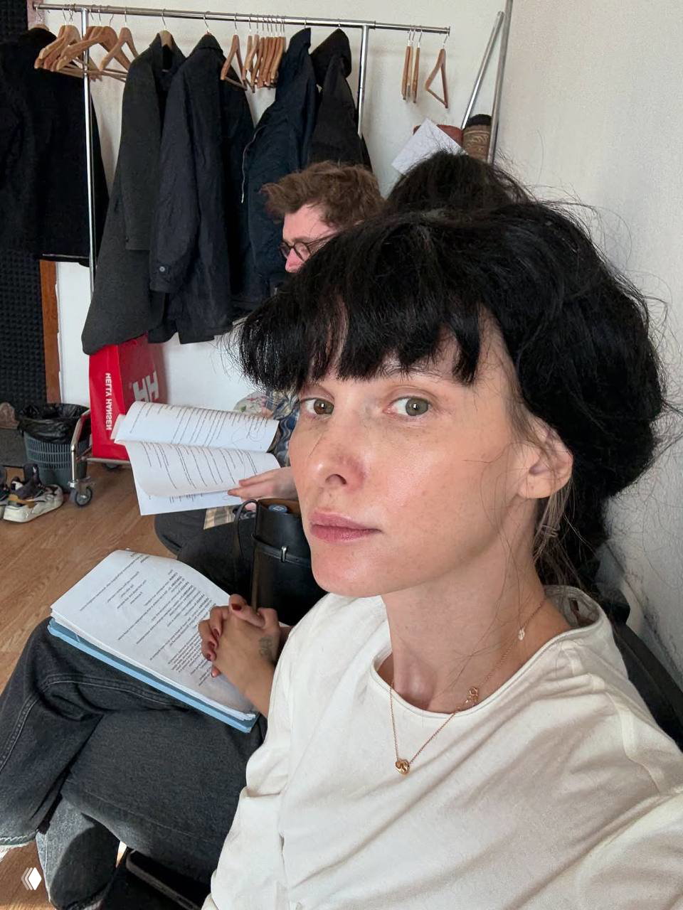 Подготовка к репетиции: актриса на фоне вешалок с одеждой держит сценарии, рядом сидит коллега, видно рабочую атмосферу.