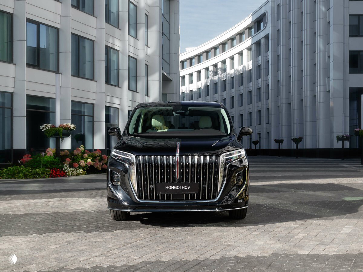 Фронтальный вид HONGQI HQ9: большая хромированная решётка радиатора и светотехника на фоне архитектуры делового квартала.