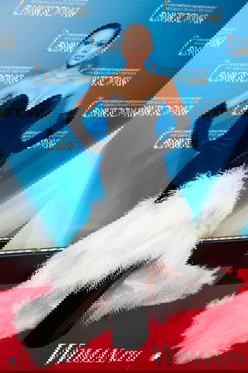 Артистка в чёрном корсетном платье с длинными перчатками и пушистым белым шлейфом позирует на фоне брендволла премии «Виктория-2026».