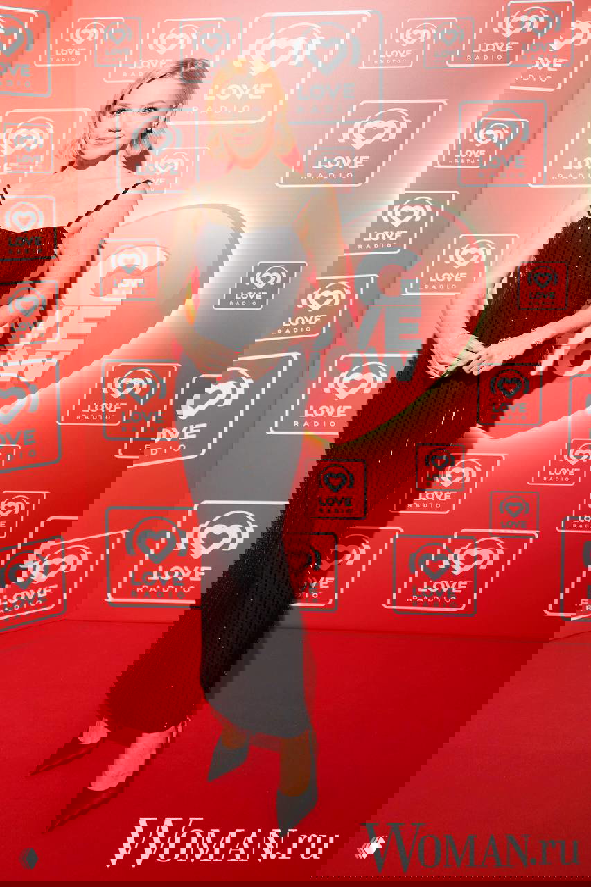 Женщина в чёрном облегающем платье в пайетках стоит на красной дорожке с логотипом Big Love Show за спиной.