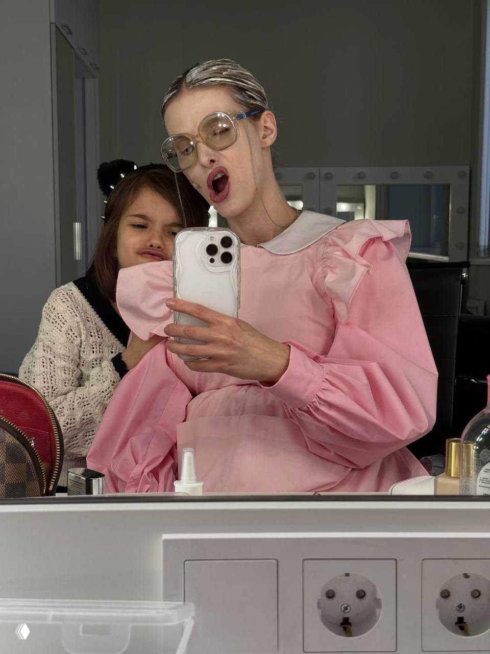 Актриса делает селфи у гримёрного зеркала вместе с ребёнком, она в розовом костюме и гримной мимике, на столе видны косметика и розетки.