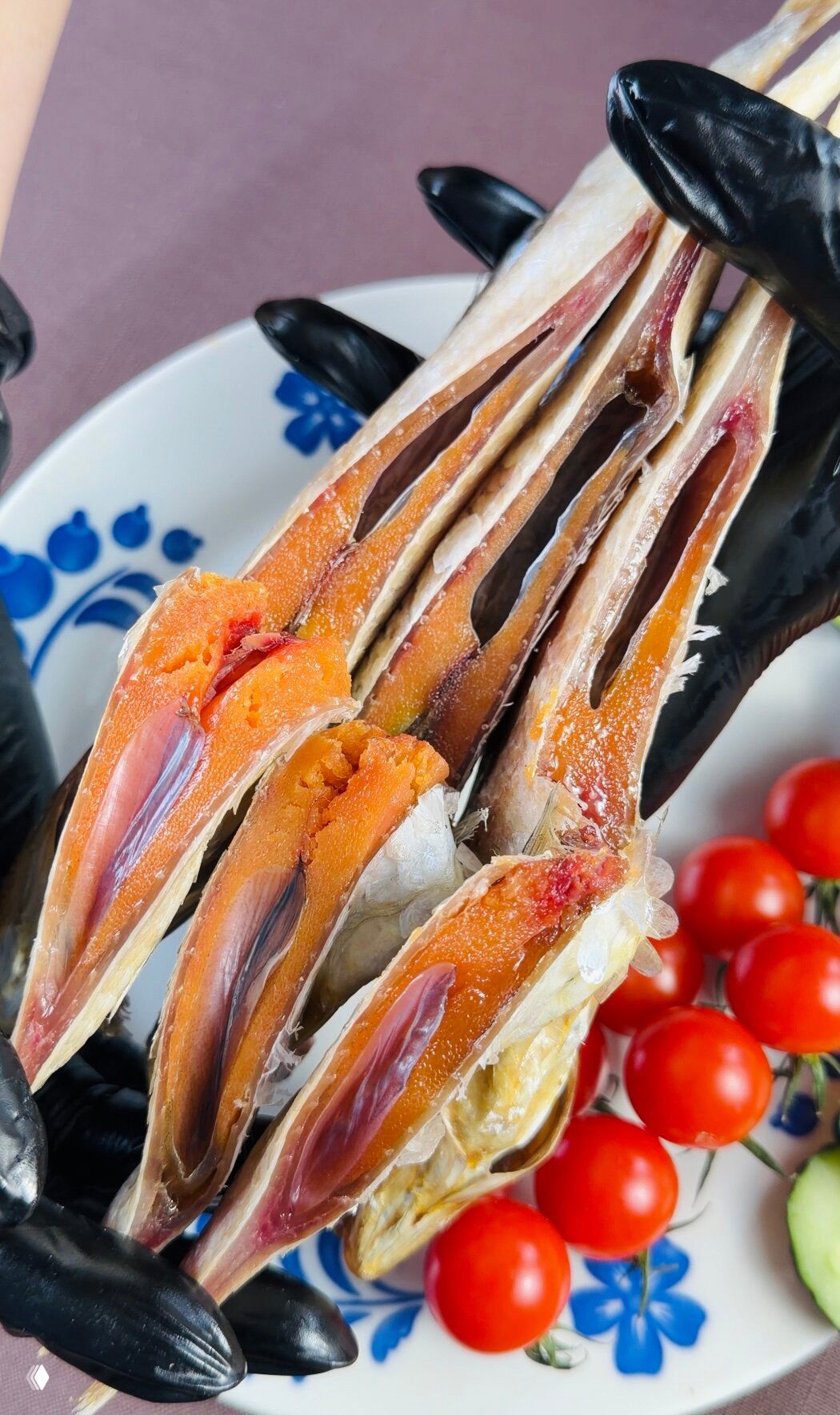 Крупный план разрезанной вяленой рыбы с яркой икрой, руки в чёрных перчатках держат филе, на фоне помидоры черри и тарелка для сервировки.