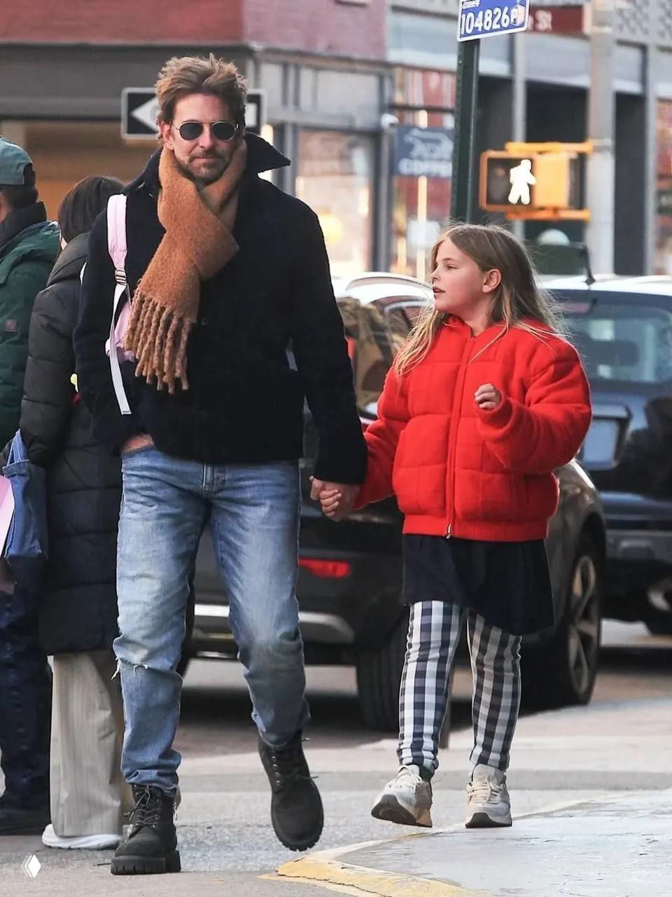 Мужчина в солнечных очках и шарфе идёт рядом с девочкой в красной куртке; уличная сцена с пешеходным переходом и машинами на заднем плане.