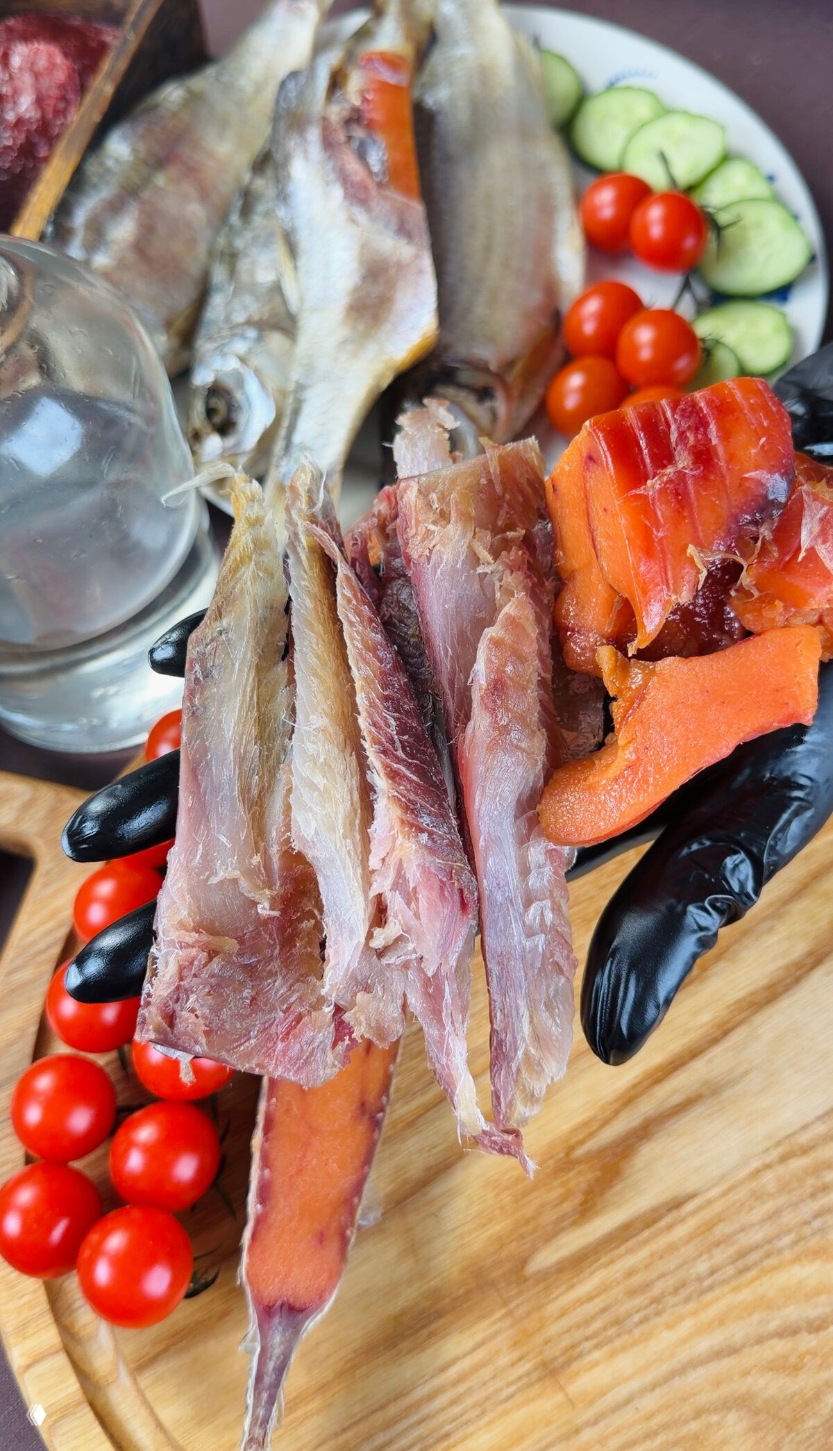 В руках в чёрных перчатках полоски вяленого филе и кусочки икры рядом с целыми рыбами и помидорами — детальный вид текстуры мяса и насыщенного цвета.