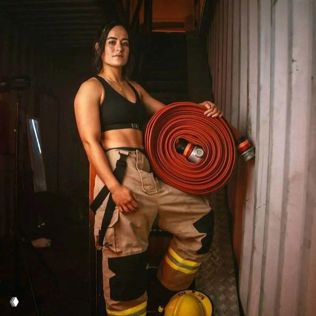 Одна женщина‑пожарный в защитных брюках держит свернутый пожарный рукав внутри ангара; поза подчёркивает силу и профессионализм.