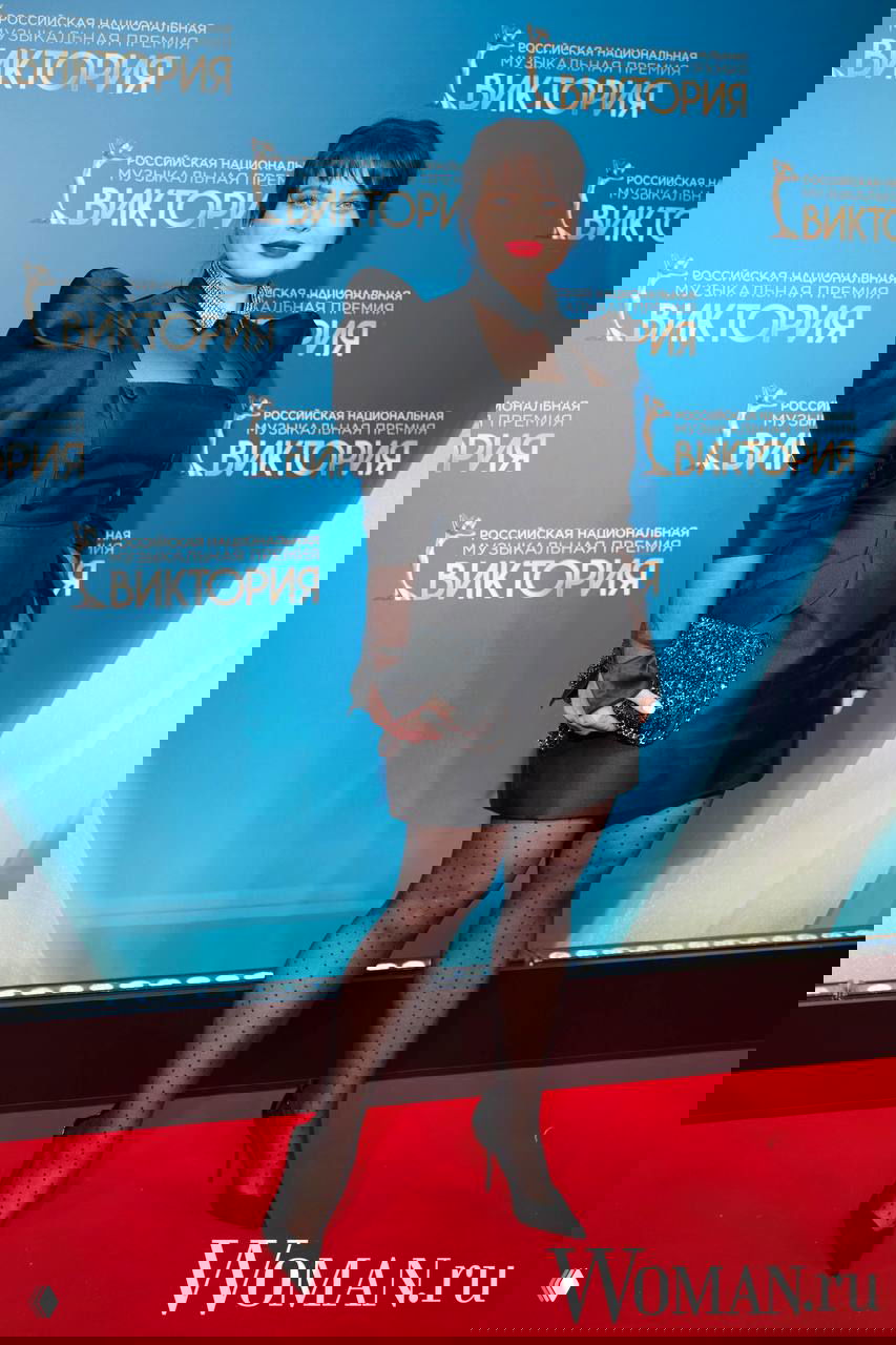 Гостья в чёрном коротком платье с пышными плечами, прозрачными колготками в горох и яркой помадой на красной дорожке премии «Виктория-2026».