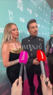 Двое артистов в вечерних нарядах дают интервью у пресс-волла на музыкальном мероприятии; в кадре несколько микрофонов, бейджи и заметный водяной знак СМИ.