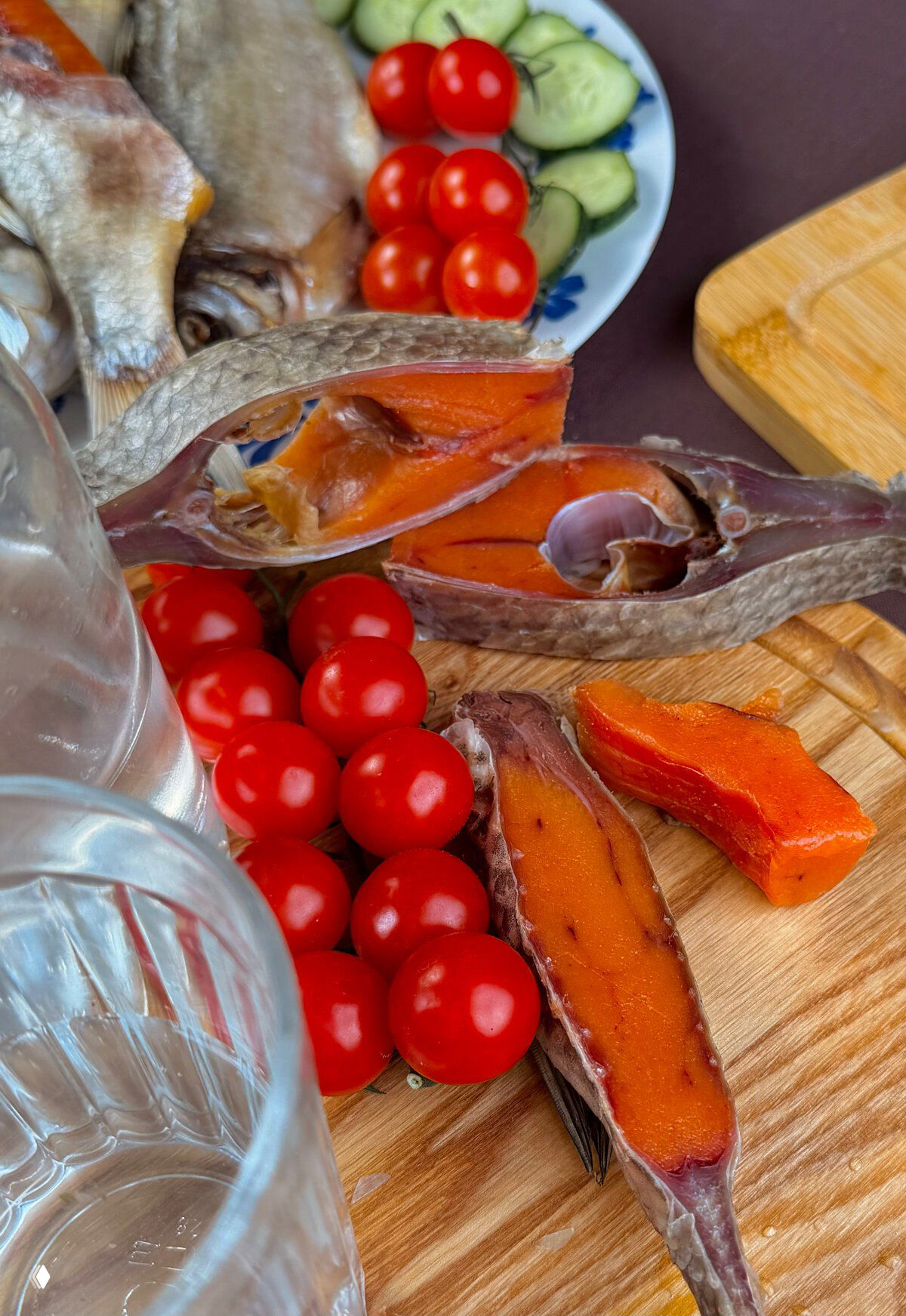 Кусочки вяленой рыбы с насыщенным оранжевым цветом икры на деревянной доске, рядом помидоры черри и кружки огурца — сервировка для подачи.