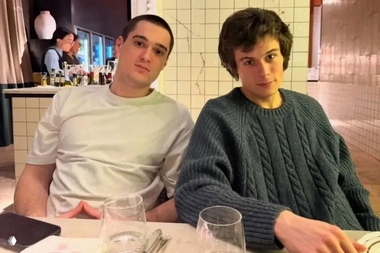 Двое молодых мужчин сидят за столом в ресторане: один в белой футболке, другой в вязаном свитере; на столе бокалы, посуда и телефон.