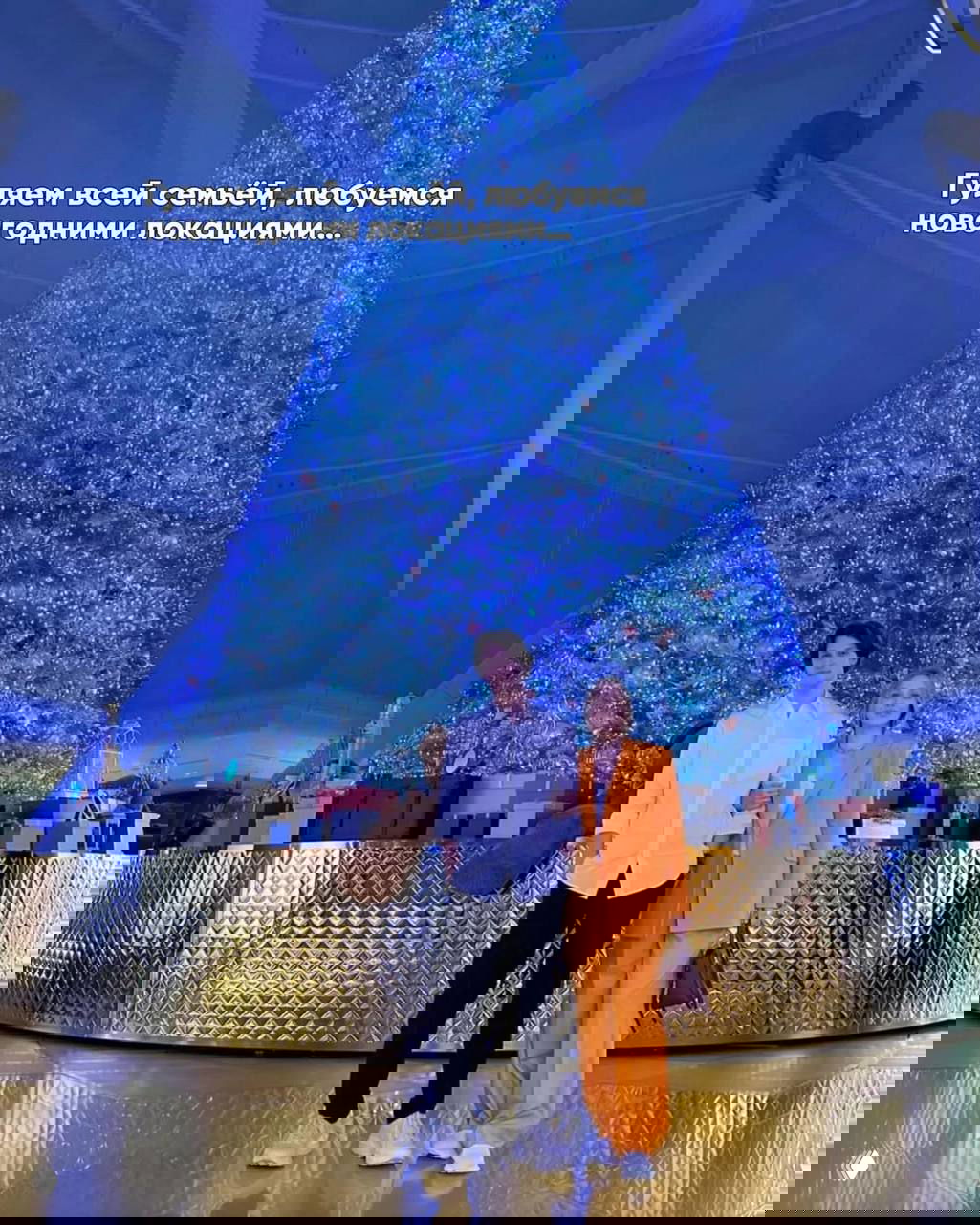 Большая голубая подсветка рождественской ёлки в публичном интерьере; на переднем плане Альбина Джанабаева со старшим сыном у огромной новогодней инсталляции.