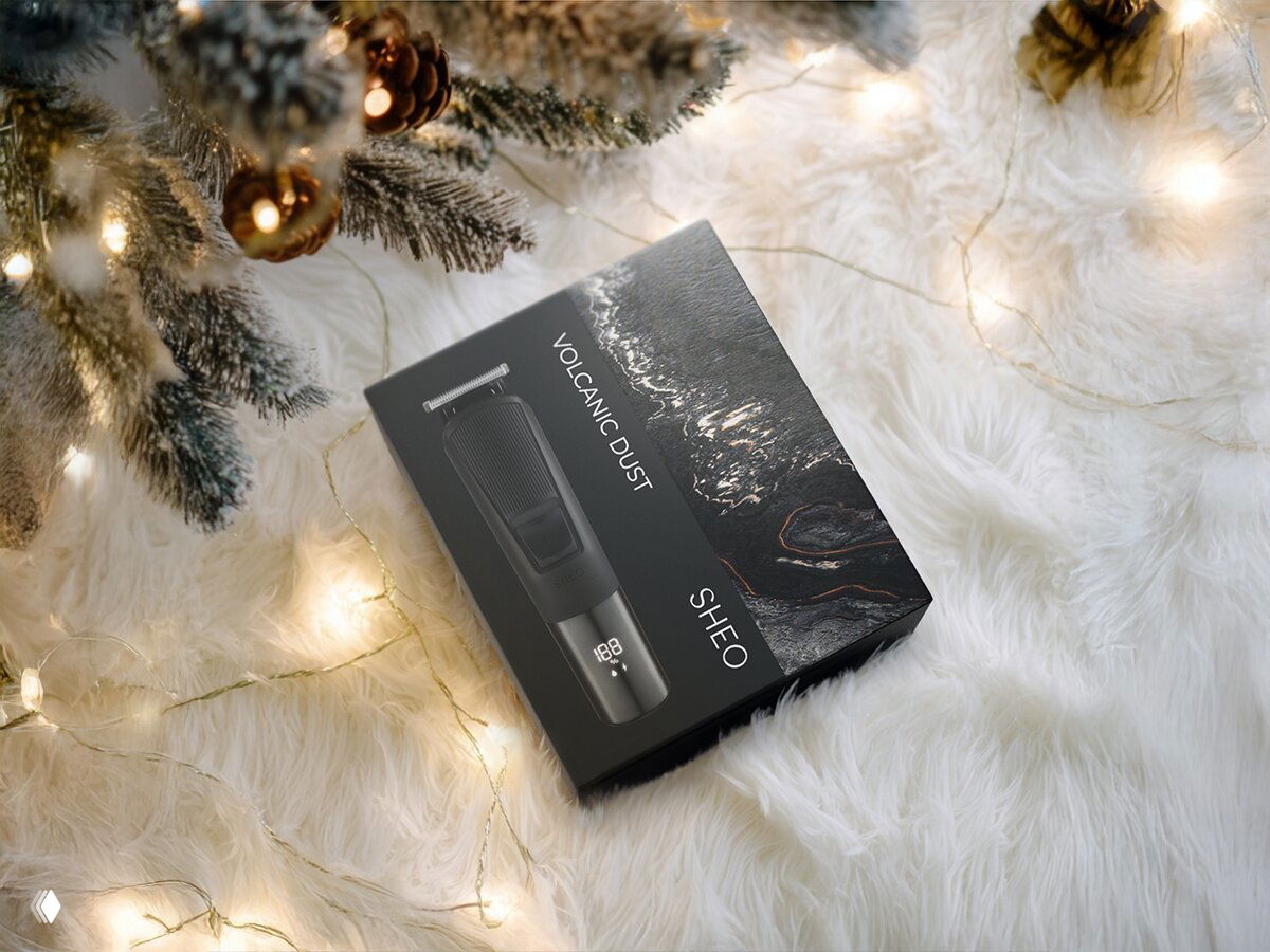 Упаковка триммера Sheo Volcanic Dust лежит на пушистом белом фоне под елочными ветками и мягкими огнями гирлянды.