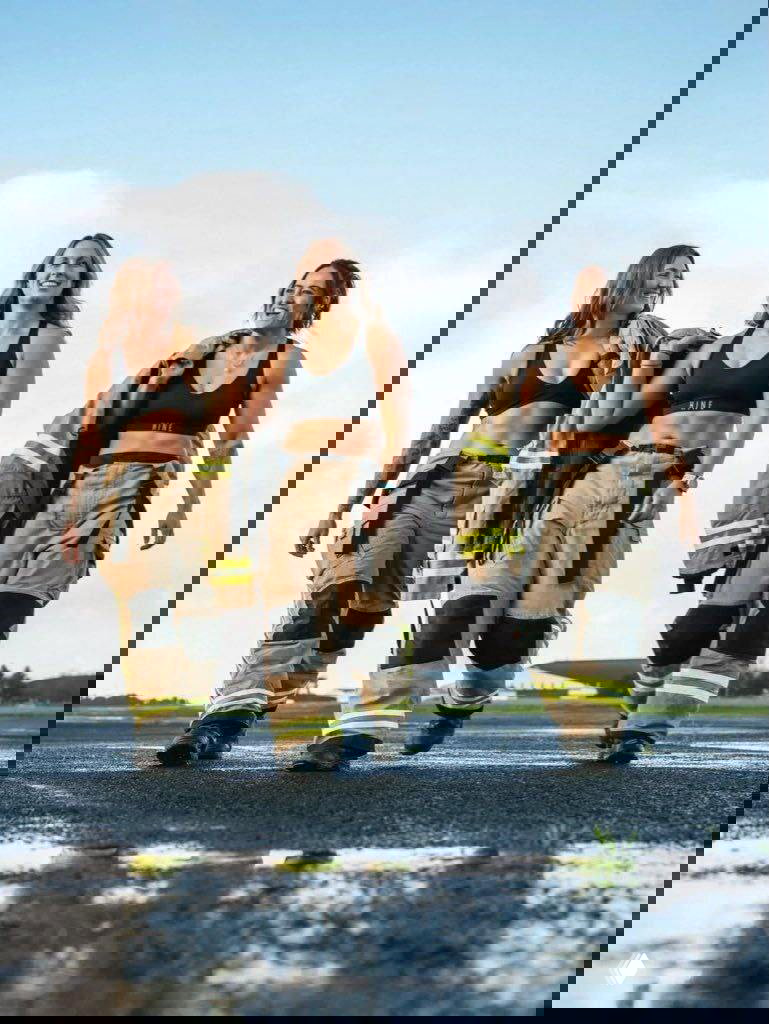 Три женщины‑пожарные идут по асфальту в брюках и коротких топах, на плечах наброшены куртки; кадр с отражением на мокрой поверхности.